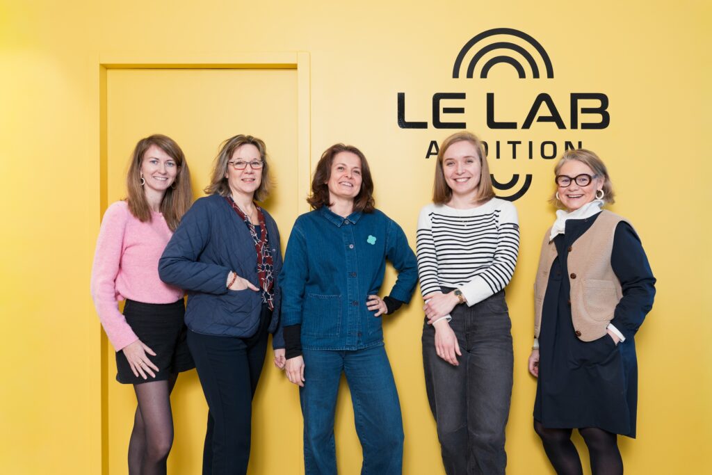 Équipe Le Lab Audition composées de 5 audioprothésistes, posant devant le logo de l'entreprise.